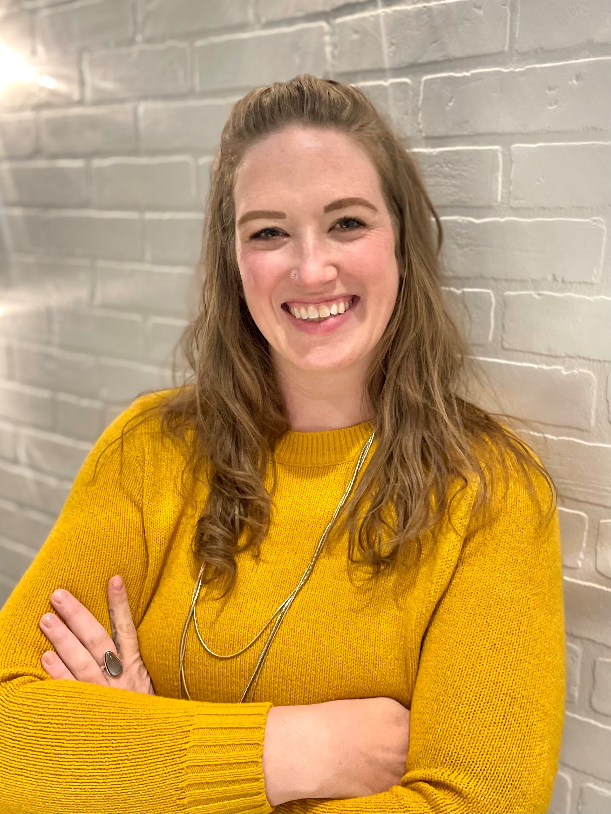 Women with long brown hear smiling with arms crossed in a yellow sweater